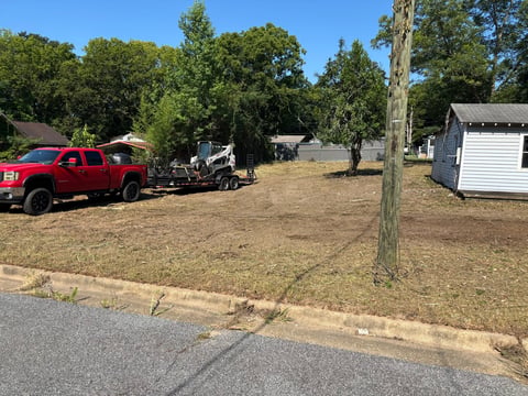 Red pickup truck parked on dirt lot with wooden utility buildings and trees under clear blue sky