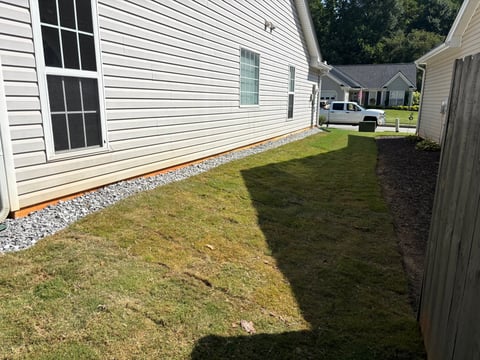 Side yard of white house with green grass lawn, gravel border, and neighboring properties visible