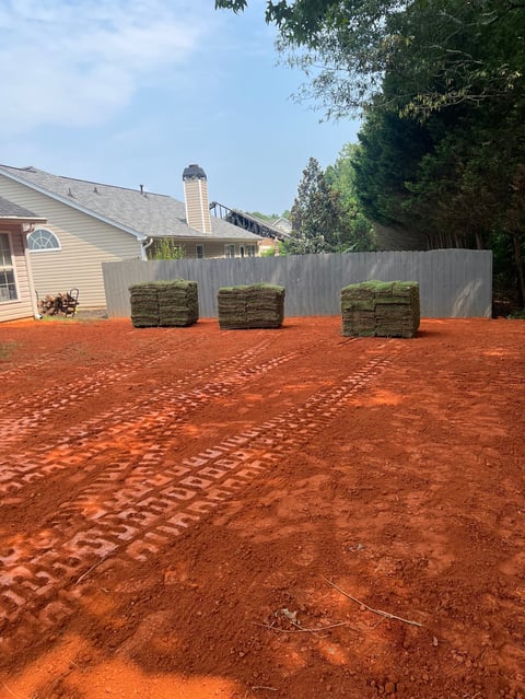 Residential backyard with red clay soil, metal fence, stacked sod rolls, and house with chimney visible in background