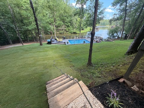 Waterfront backyard with wooden dock leading to blue boat, manicured lawn, towering trees, and calm river in background