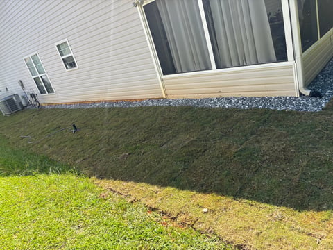 Sloped backyard with patchy lawn beside a white house foundation and gravel border