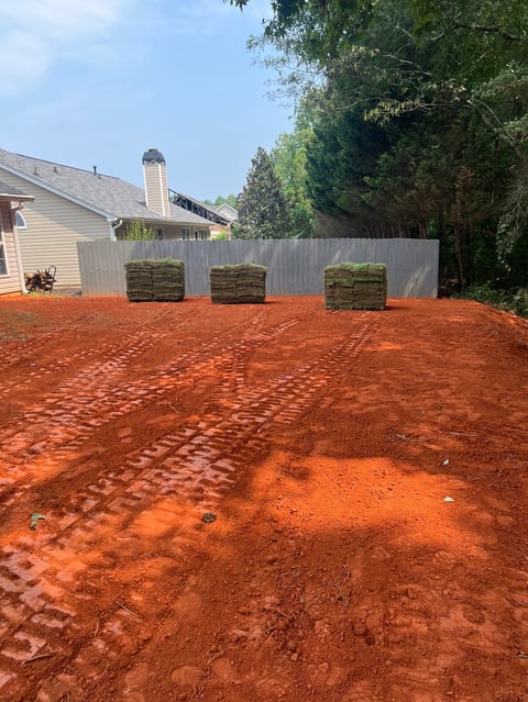 Residential yard with reddish clay soil, white corrugated metal fence, and stacked sod rolls. House and trees visible in background.
