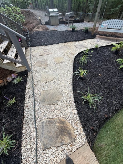 Aerial view of a backyard with stone pathway, mulched garden beds with plants, grill, and forest surrounding the property