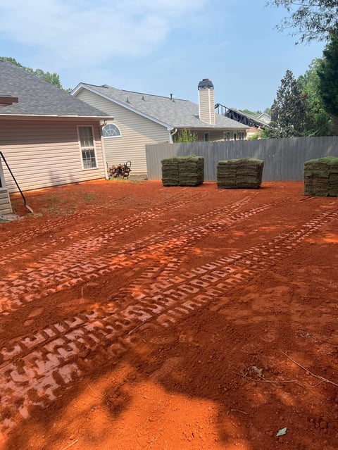 Residential backyard with freshly laid red clay or dirt, stacked sod rolls, and a beige two-story house with gray roof in background
