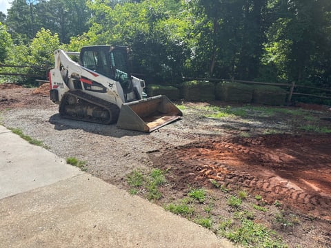 White skid steer loader with red accents working on dirt ground in a wooded area next to a residential property