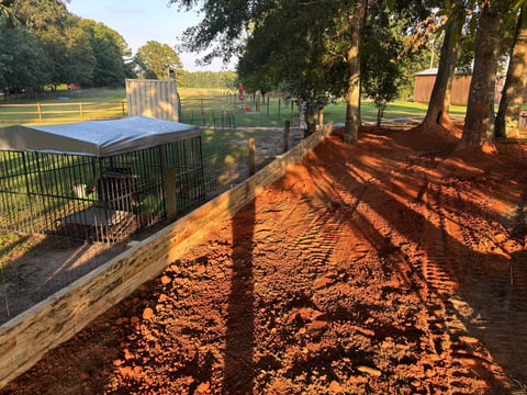 A rural farm property with livestock pens and fencing in a sunny field surrounded by large oak trees casting shadows on red dirt ground