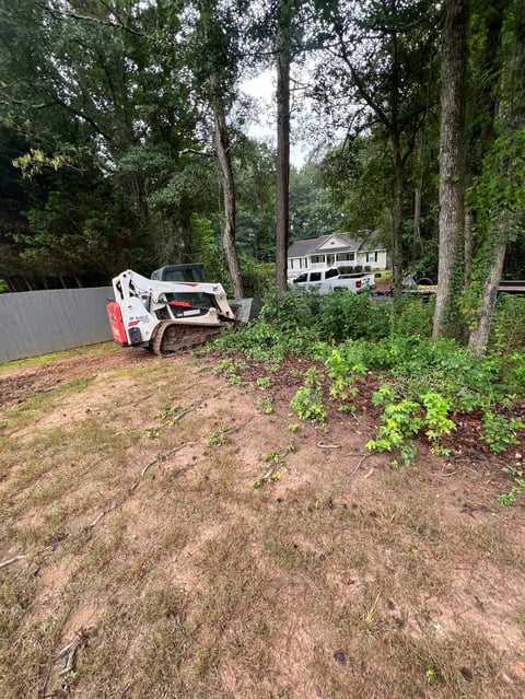 White and red Bobcat skid-steer loader on cleared land surrounded by tall trees with a house visible in the background