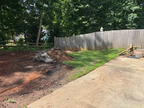 Backyard landscape with wooden fence, mulched garden bed on left, concrete patio on right, and scattered construction materials