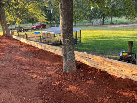 Park playground with red mulch border, wooden log edging, large tree, and grass field with playground equipment in background