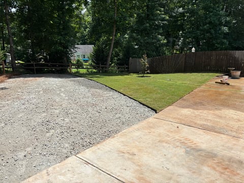 Backyard with gravel driveway, manicured lawn, wooden deck, and tall fence surrounded by trees