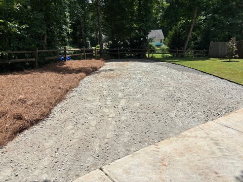 Gravel driveway under construction with mulch and grass alongside, bordered by trees and a fence