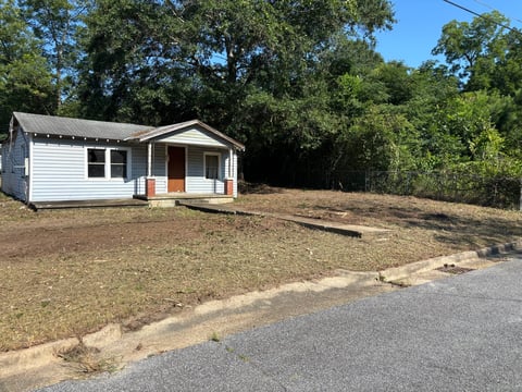 Small white single-story house with covered porch on sparse yard surrounded by trees