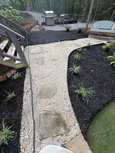 Overhead view of backyard landscape featuring gravel pathways, planted ornamental grasses, and outdoor grill area surrounded by tall trees