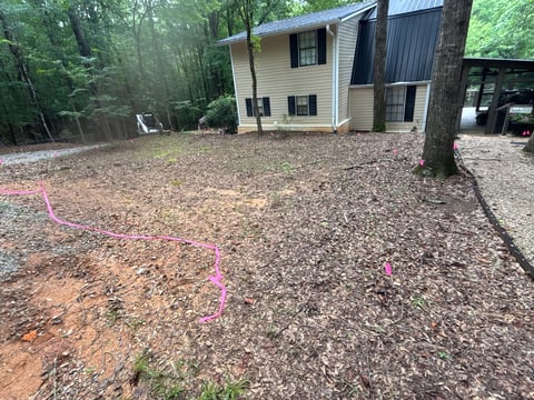 Two-story residential house with beige siding and metal roof on wooded lot with pink survey flags in dirt yard