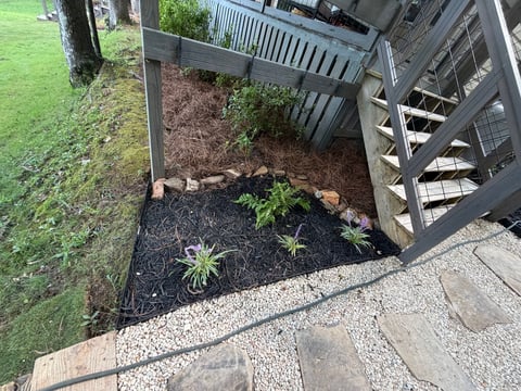 Overhead view of a residential deck corner with landscaping bed, young plants, mulch, gravel pathway, and adjacent green lawn