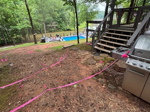 Backyard deck construction site with pink marked utility lines, wooden stairs, pool, and outdoor grill surrounded by trees